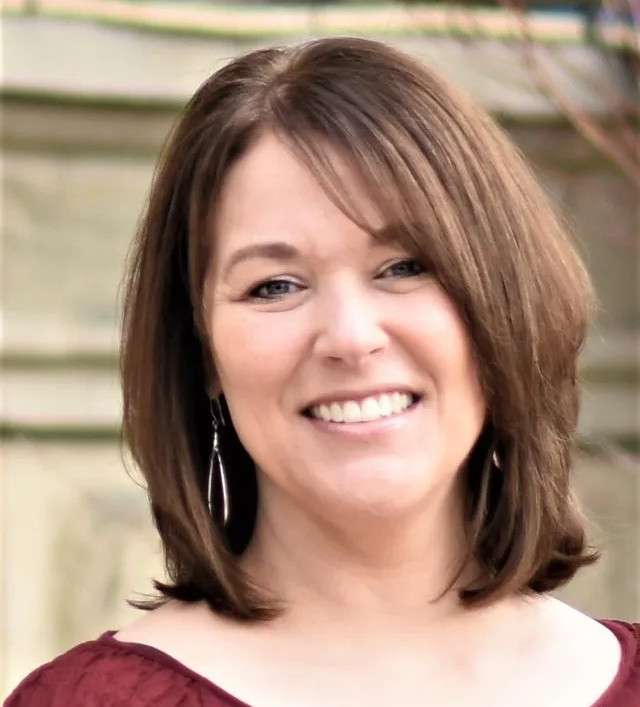 A woman with shoulder-length brown hair, wearing a maroon top and silver drop earrings, smiles at the camera in an outdoor setting with a blurred background.