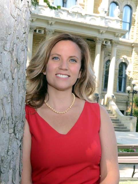 Lindsey-Website A woman with wavy blonde hair, wearing a red sleeveless dress and a pearl necklace, stands beside a tree. Behind her is a historic building with arched windows and stone steps. She is smiling at the camera.