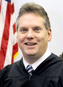 JUDGE_PADDEN_PIC-219×300 A smiling man in a black judge’s robe and striped tie stands in front of an American flag.