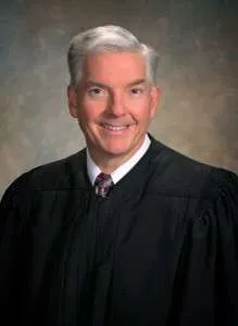 Judge-David-Bennett-close-240×300 A middle-aged man with short gray hair wearing a black judges robe, white shirt, and patterned tie, smiling at the camera against a neutral, softly blurred background.