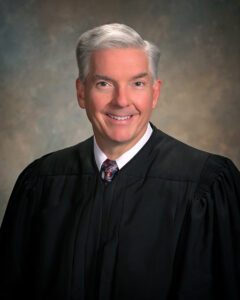 Judge-David-Bennett-close-240×300 A middle-aged man with short gray hair wearing a black judges robe, white shirt, and patterned tie, smiling at the camera against a neutral, softly blurred background.