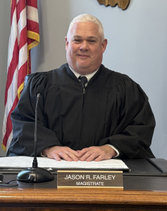 Magistrate-Farley-2025-237×300 A man in a black judge’s robe sits at a desk with a nameplate reading “Jason R. Farley, Magistrate.” An American flag is visible in the background.