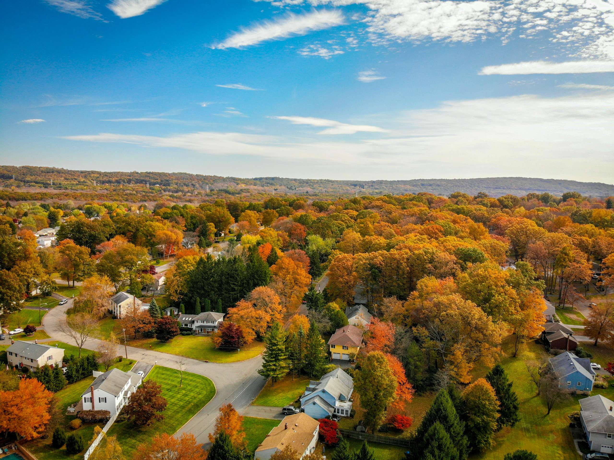 Aerial view of a suburban neighborhood surrounded by trees with autumn foliage in shades of orange, yellow, and green under a partly cloudy sky. Houses are spread along curved roads with lawns and driveways.