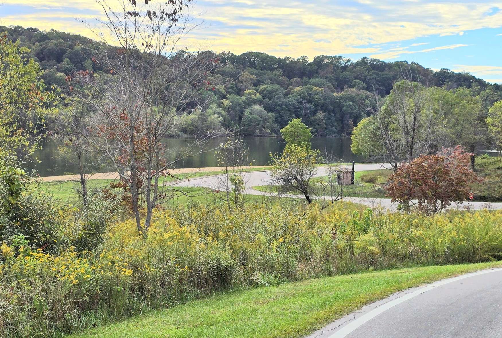 A paved road curves through a park with green grass, wildflowers, trees, and a lake in the background. The sky is partly cloudy, with forested hills lining the horizon.