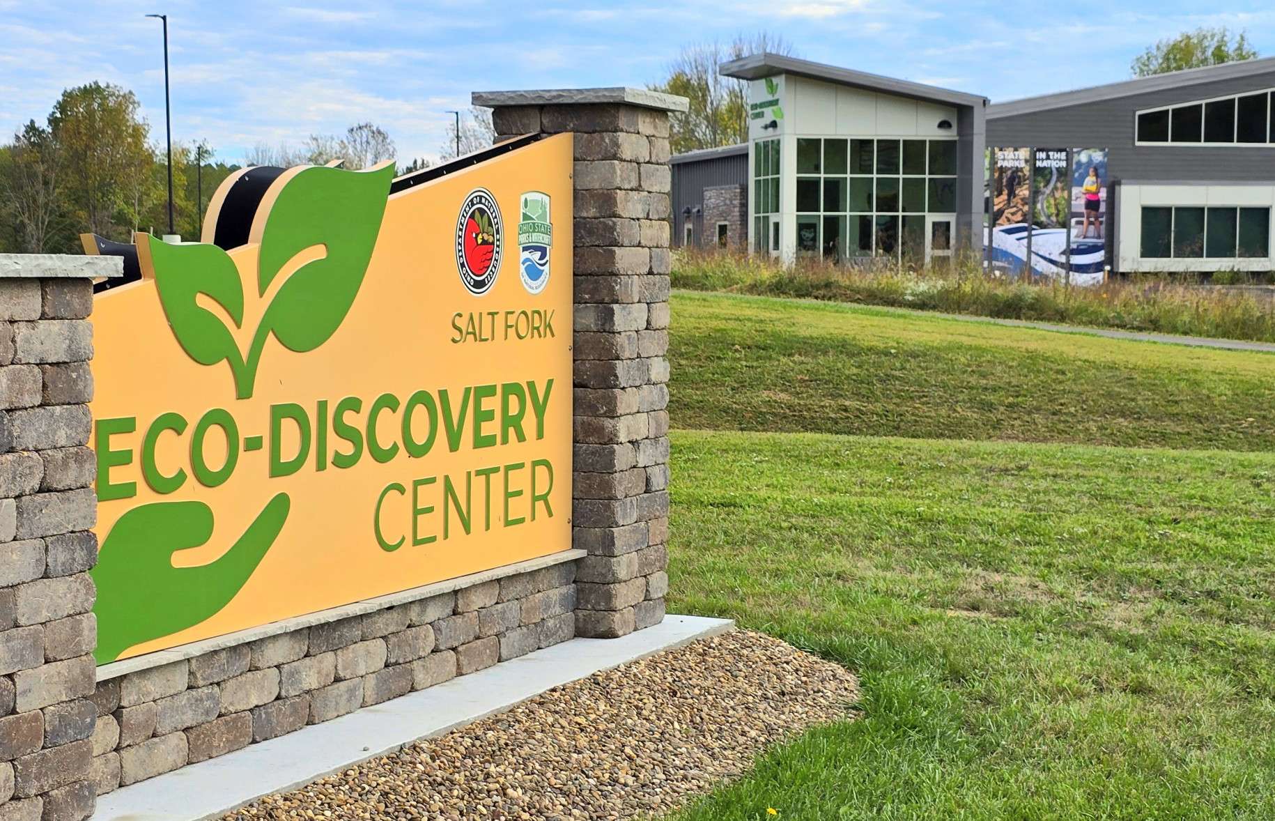 A large yellow sign reading Eco-Discovery Center stands on a grassy lawn in front of a modern building with large windows. The sign also features the Salt Fork logo and green leaf graphics. Trees are visible in the background.