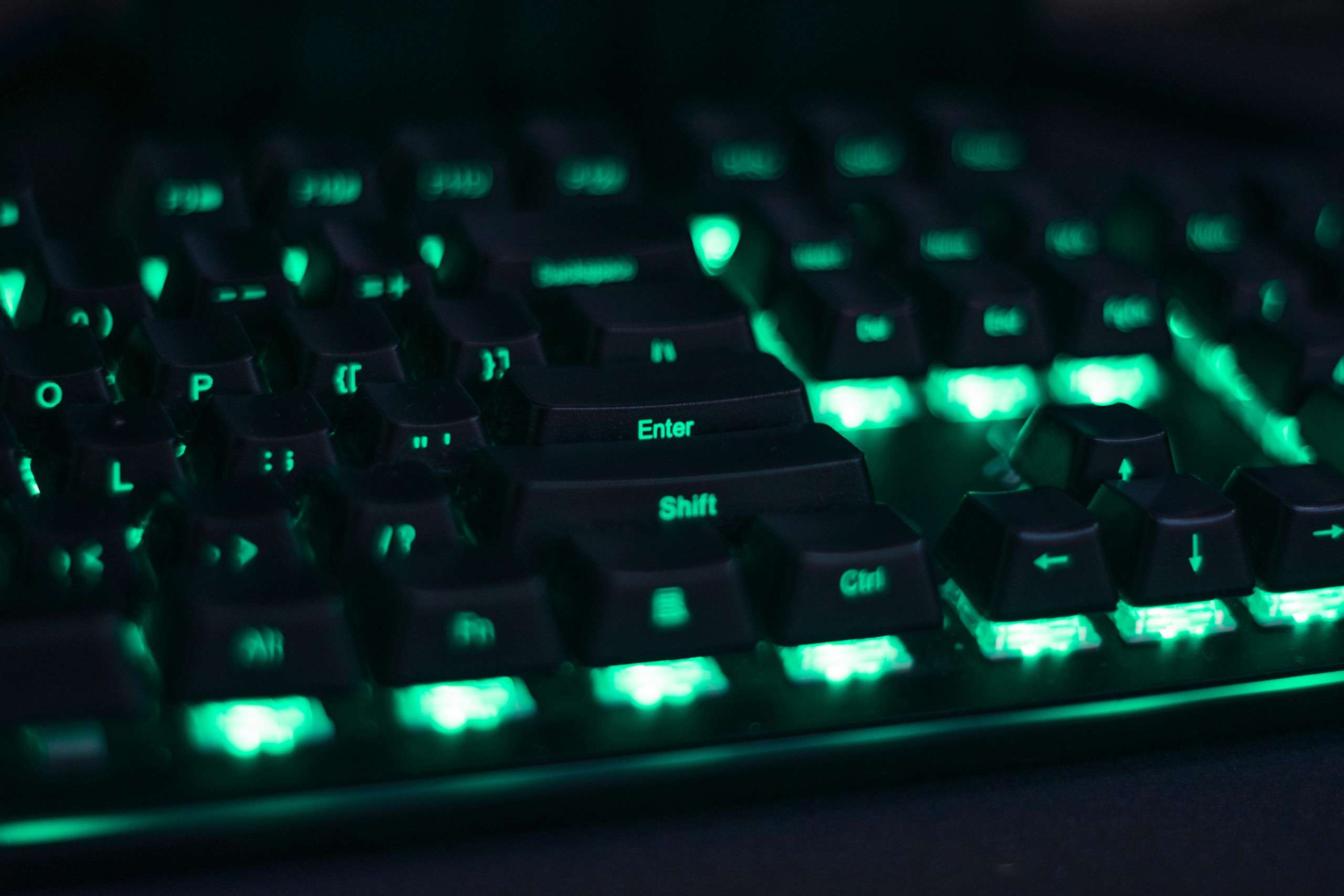 A close-up of a black keyboard with teal backlighting, highlighting the Enter and Shift keys. The glowing keys create a futuristic and vibrant look in a dark environment.